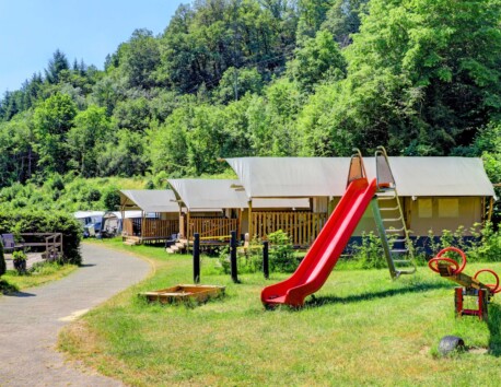 Spielplatz mit dahinter liegenden Safarizelten und Stellplätzen an einem bewaldeten Hügel auf dem Campingplatz Drei Spatzen