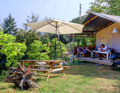 Vakantiegangers relaxen op veranda safaritent met ervoor picknicktafel en parasol Camping Pian d'Amora